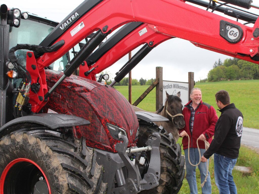 Doi bărbați vorbesc lângă un tractor Valtra și un cal pe un drum de pământ.