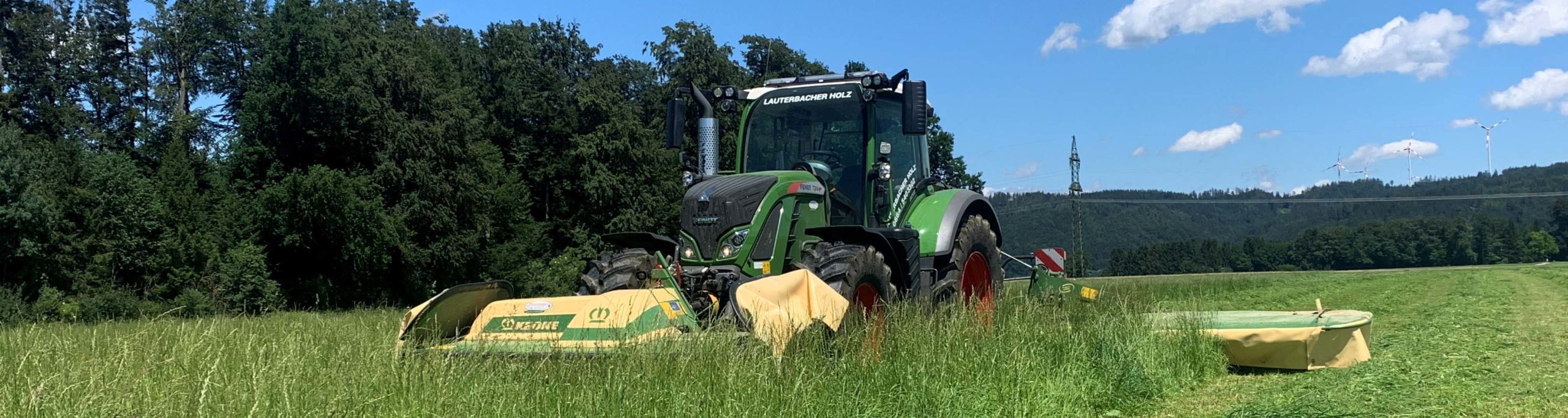 Grüner Fendt-Traktor mäht Gras auf einem Feld mit Wald im Hintergrund.