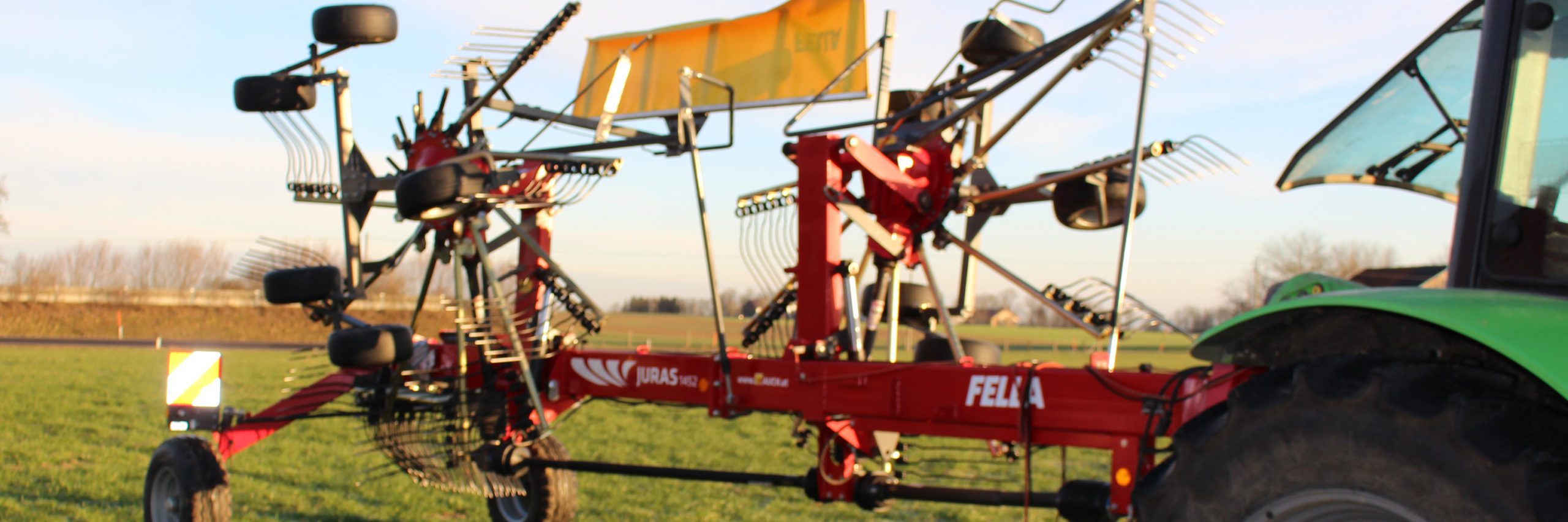 Landwirtschaftliches Gerät FELLA JURAS 1452 neben einem grünen Traktor auf einem Feld.