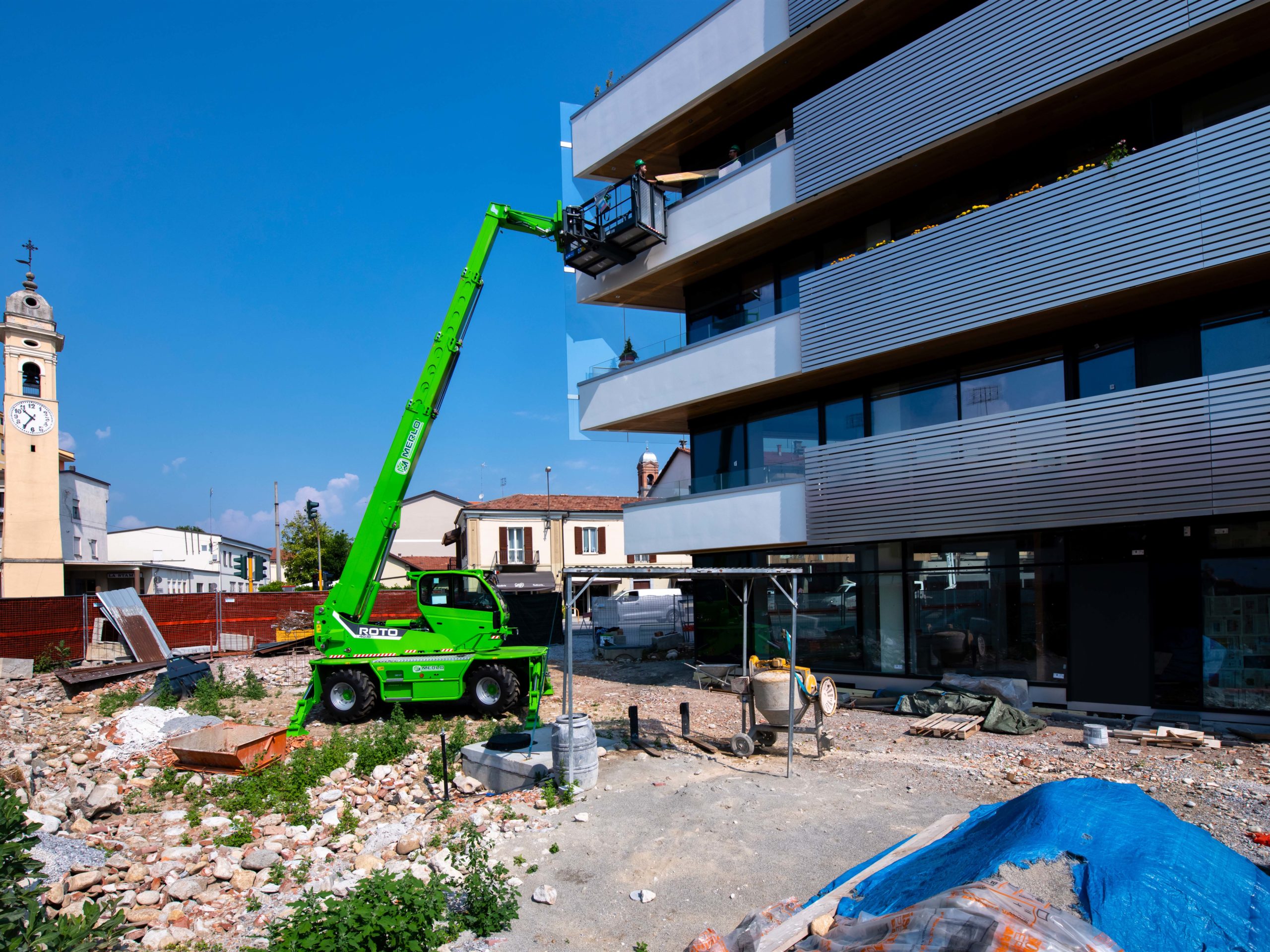 Ein grüner Merlo ROTO 40.18 Kran hebt Material zu einer Baustelle an einem modernen Gebäude.