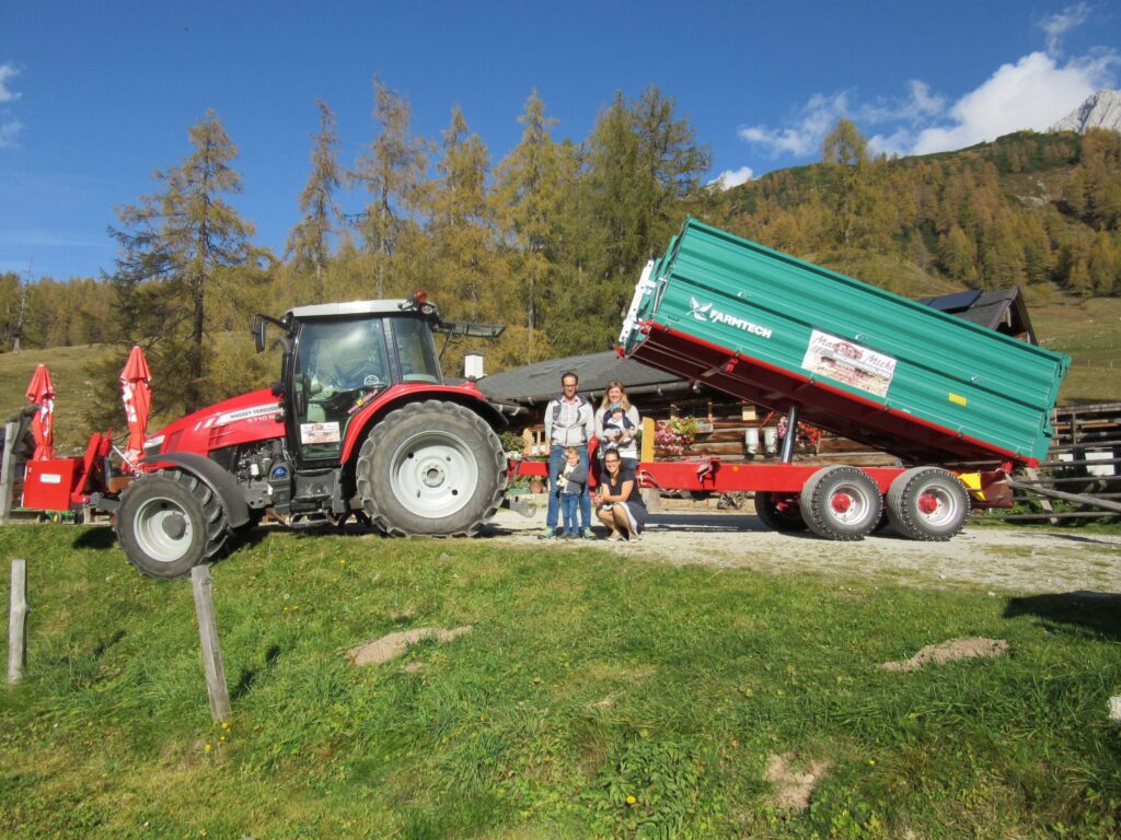 Ein roter Traktor von Massey Ferguson zieht einen grünen Farmtech-Anhänger, daneben steht eine Gruppe von fünf Personen.