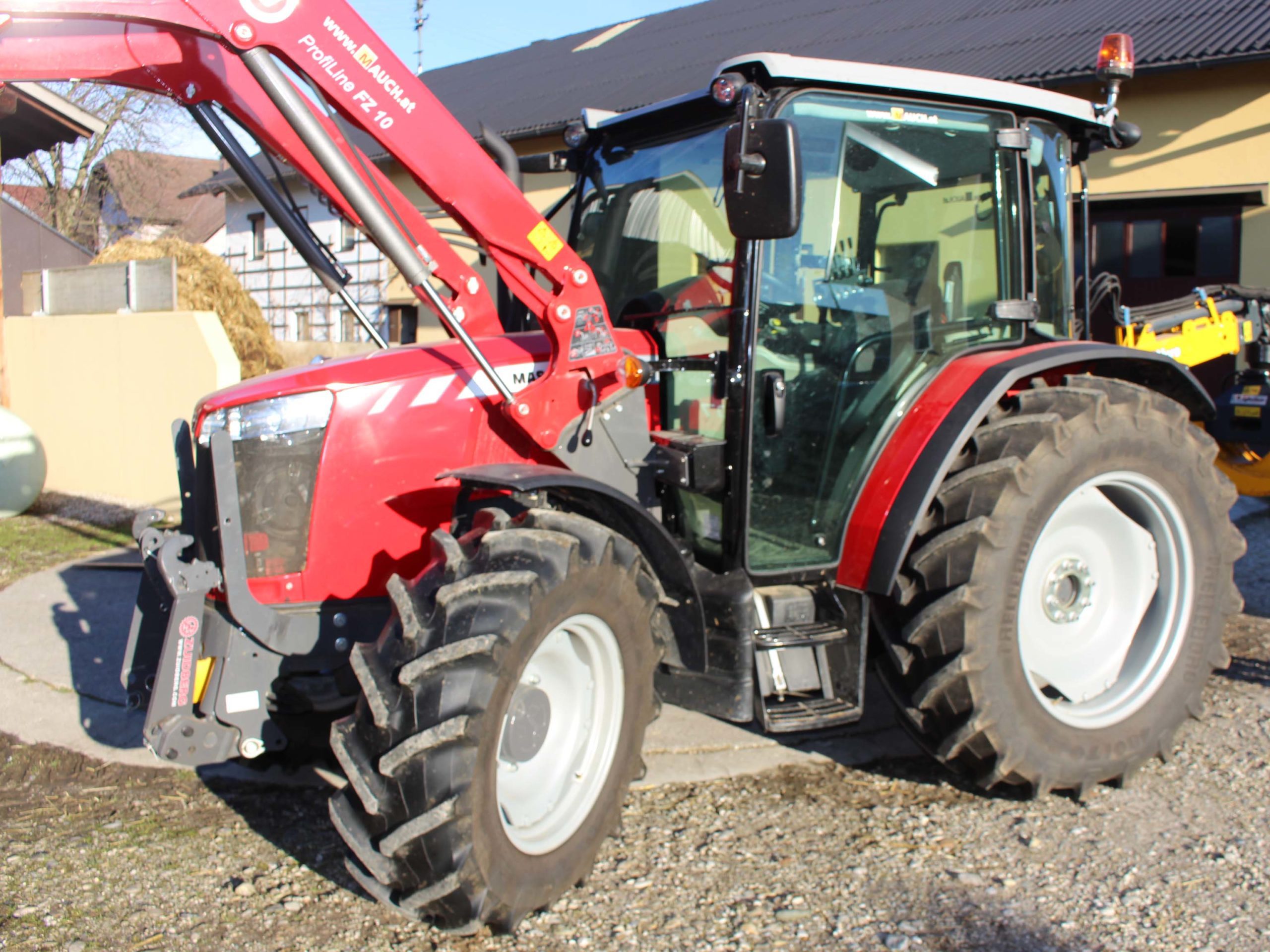 Tractor Massey Ferguson roșu cu încărcător frontal Proline FZ 10, parcat într-o curte din fața clădirilor.
