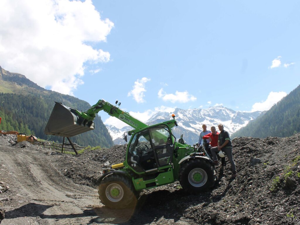 Мерло Мултифармер 40.9 са кашиком на градилишту испред планина прекривених снегом, тројица мушкараца поред њега.