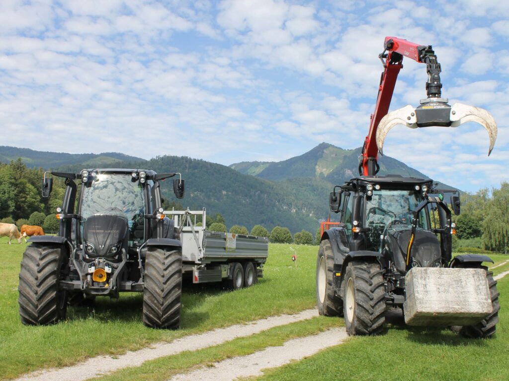 Două tractoare Valtra N154, unul cu braț de graifer și bloc de beton, pe o pajiște alpină în fața unui peisaj montan.