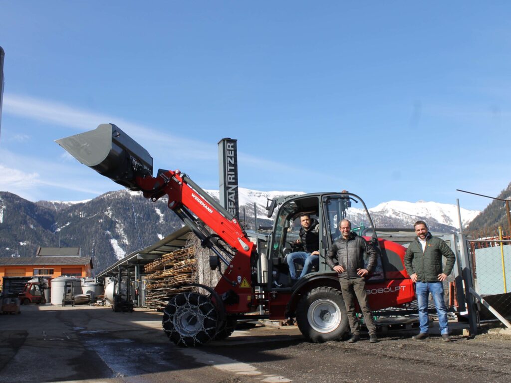 Тројица мушкараца стоје испред утоваривача на точковима Вајдеман ВМ3080 ЛПТ са ланцима за снег, у позадини планине прекривене снегом.