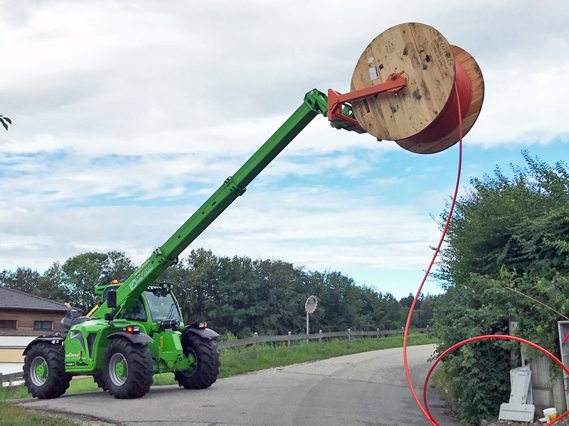 Ein grüner Merlo MF 40.9 CS-170 CV Tronic hebt eine große Kabeltrommel auf einer Straße.