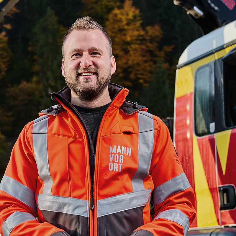 Ein Mann in einer orangefarbenen Jacke mit der Aufschrift Mann vor Ort steht vor einem Lieferfahrzeug.