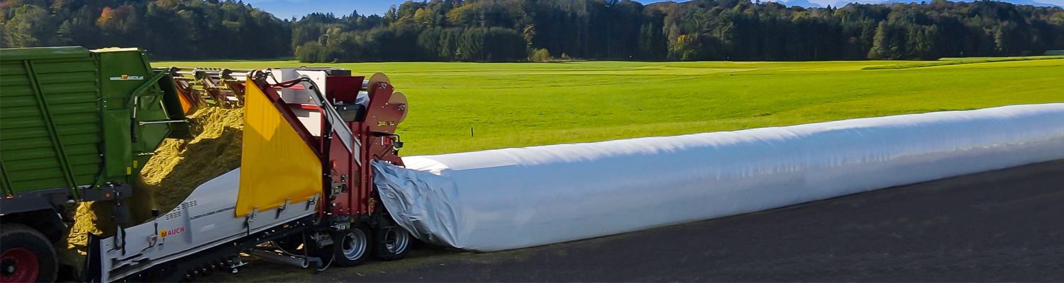 Ein Silospeed Befüllsystem verpackt Silage auf einem flachen Feld mit grünen Wiesen und Wald im Hintergrund.