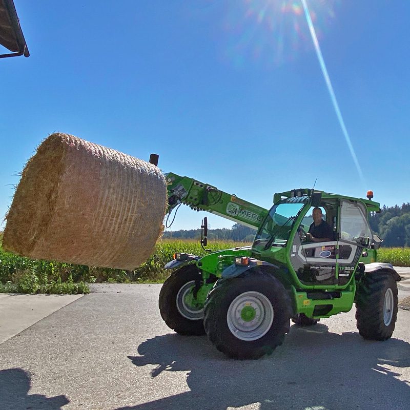 Ein Merlo TF38.10 Teleskoplader hebt eine große Heuballenrolle an.