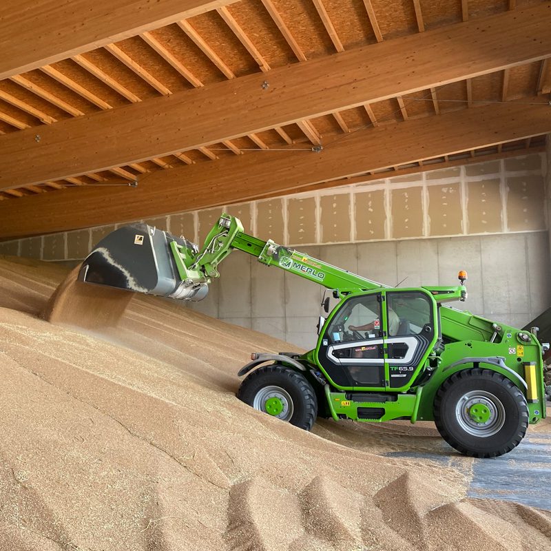 Grüner Merlo P65.9 CS Teleskoplader schaufelt Getreide in einer Lagerhalle.