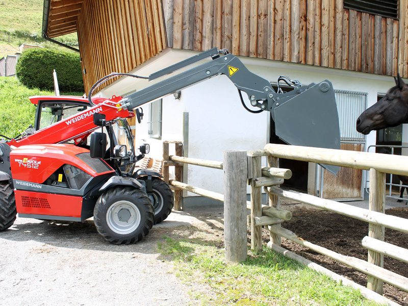 Ein Weidemann T4512 Teleskoplader hebt eine Schaufel vor einem Bauernhof an.