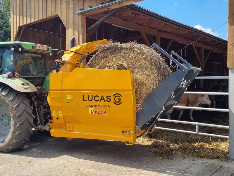 Ein Traktor mit einem gelben Lucas CASTOR+ 20R Futtermischwagen, der Heu in einen Stall transportiert.