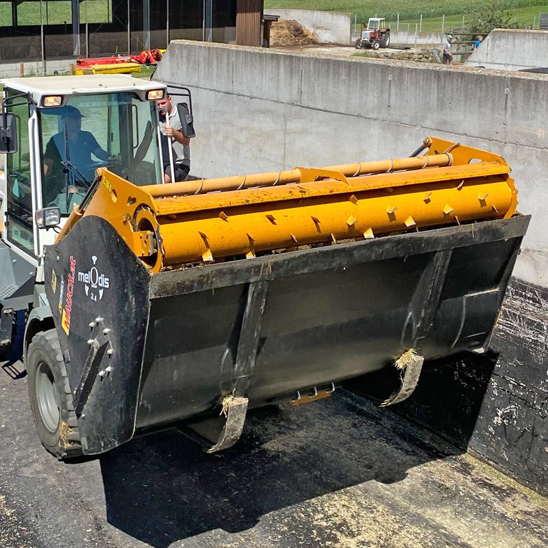 Traktor mit Melodis 2.4 Mähwerk, der Heu in eine Betonhalle transportiert.