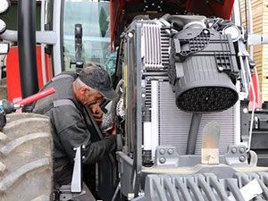 Un bărbat în jachetă de lucru roșie stă în fața unui autobuz decapotabil într-o zonă rurală.
